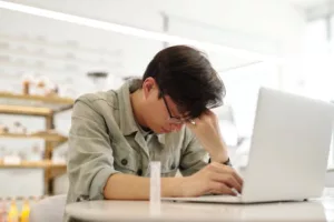 Exhausted young man working on a laptop inside a bright room, displaying fatigue.