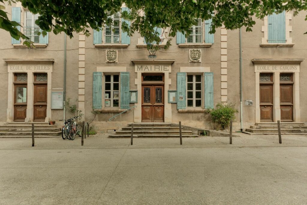Façade d'une mairie de village en pierre, avec volets bleus, sous des arbres en été