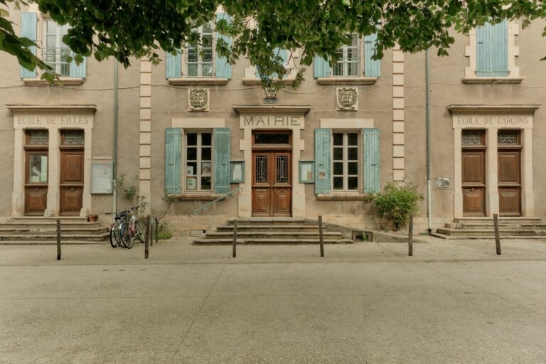 Façade d'une mairie de village en pierre, avec volets bleus, sous des arbres en été